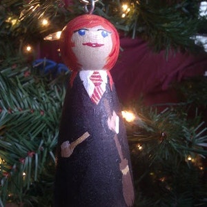 May include: A hand-painted wooden Christmas ornament depicting a character with red hair, blue eyes, and a black robe. The ornament has a silver loop for hanging and features a broom and a wand.