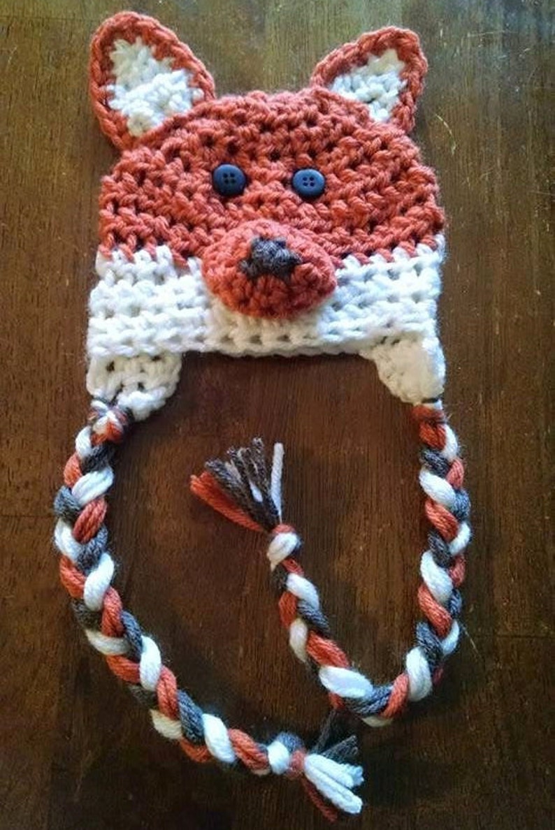 以下が含まれることがあります： オレンジと白の糸で編まれたキツネの帽子。帽子にはオレンジ、白、グレーの糸で編まれた三つ編みの尻尾が付いている。
