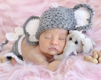May include: A newborn baby wearing a gray and white crocheted elephant hat and a matching outfit, sleeping on a pink fuzzy blanket with a gray elephant plush toy.