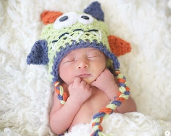 May include: A sleeping baby wearing a green, blue, and orange crocheted monster hat. The hat has a large eye and a stitched-on mouth.