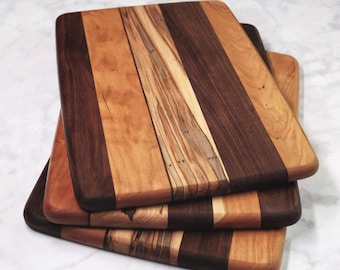 May include: A stack of rectangular wooden cutting boards. Each board features a unique pattern of alternating wood grain stripes in shades of brown, tan, and light brown. The boards have rounded corners and a smooth finish, showcasing the natural wood tones.