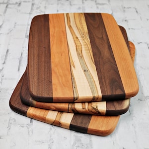 May include: Three wooden cutting boards stacked on top of each other. Each board has a rectangular shape with rounded corners and is made of different wood types, creating a striped pattern in shades of brown, tan, and cream.