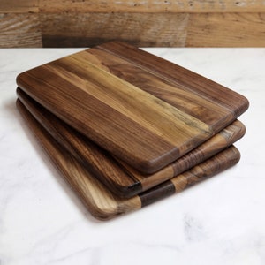 May include: A stack of three rectangular wooden cutting boards. The boards are made of dark brown wood with lighter brown and tan wood grain patterns. The edges are rounded. The boards are stacked on top of each other.