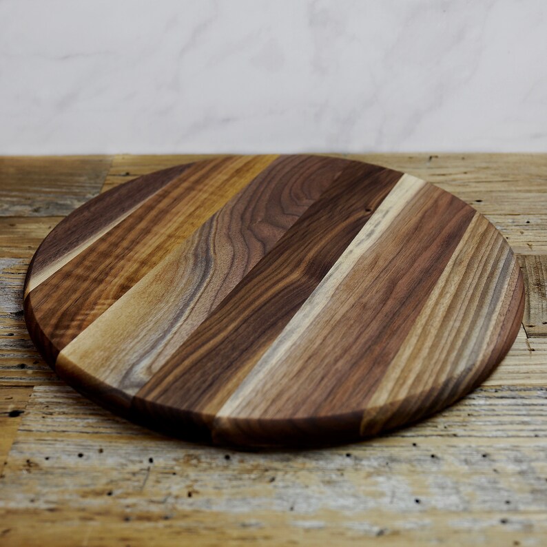May include: A round wooden cutting board made of walnut wood with a natural wood finish. The board has a diameter of approximately 12 inches.