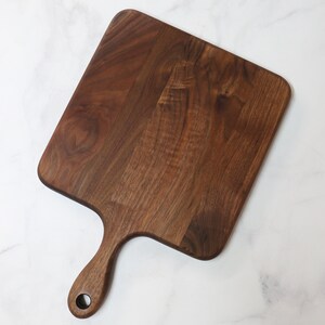 May include: A dark brown wooden cutting board with a handle. The board is square shaped and has a smooth surface.