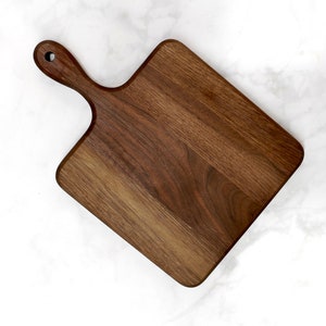 May include: A rectangular walnut wood cutting board with a handle. The board has a natural wood grain pattern and is approximately 12 inches by 16 inches.
