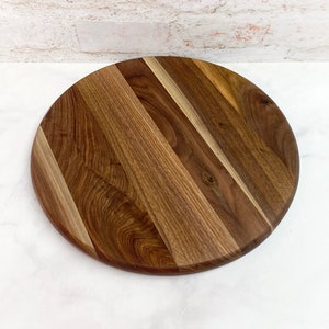 May include: Round wooden cutting board with a natural wood grain pattern. The board is made of walnut wood and has a light brown color.