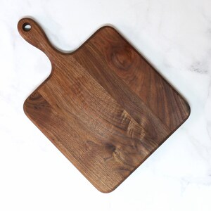 May include: A square-shaped wooden cutting board with a handle. The board is made of dark brown walnut wood and has a natural wood grain.