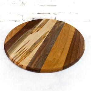 May include: Round wooden serving board with a multi-wood design. The board is made of walnut, maple, and cherry wood.