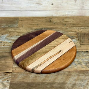 May include: A round wooden cutting board with a variety of wood tones, including light and dark browns, tan, and purple. The wood pieces are arranged in diagonal stripes. The board is approximately 12 inches in diameter.