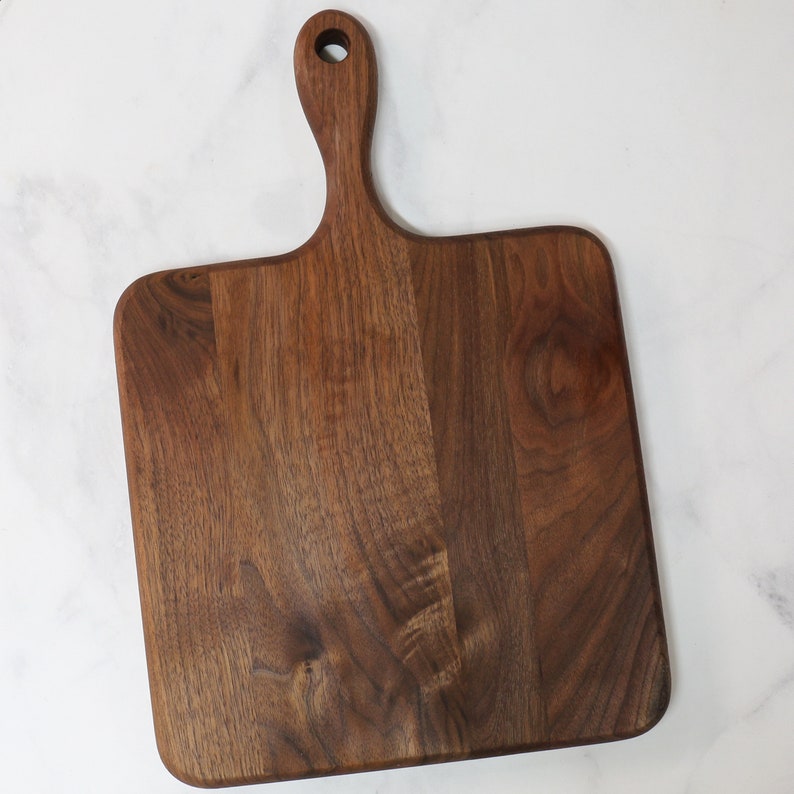 May include: A large, rectangular walnut wood cutting board with a handle. The board has a natural wood finish and is perfect for serving cheese, charcuterie, or other appetizers.