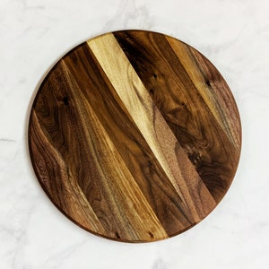 May include: A round wooden serving board with a natural wood grain pattern. The board is made of walnut wood and has a dark brown color.