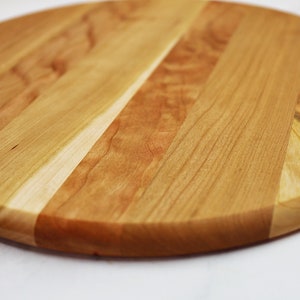 May include: A round wooden cutting board made of cherry wood with a natural finish. The board has a light brown color with darker brown streaks throughout. The board is made of several pieces of wood that have been glued together to create a unique pattern.