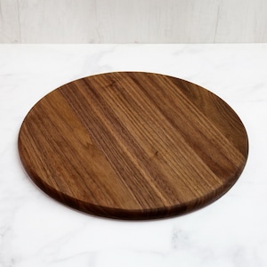 May include: A round, wooden cutting board with a rich brown color and visible wood grain. The board is circular and appears to be made of a solid piece of wood. The surface is smooth and the edges are rounded.