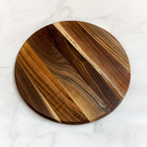 May include: Round wooden cutting board made of walnut wood with a natural wood finish. The board has a unique grain pattern and is perfect for serving cheese, charcuterie, or other appetizers.