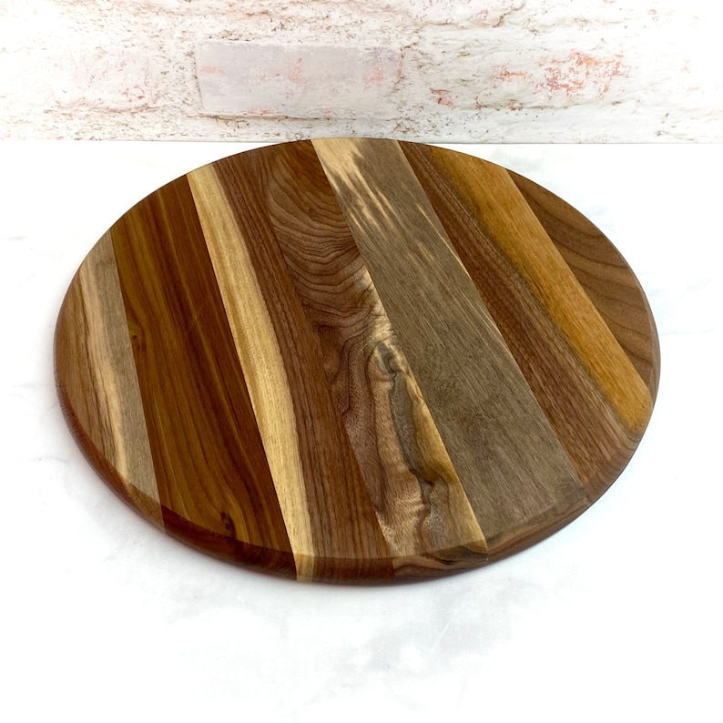 May include: A round wooden cutting board with a natural wood finish. The board is made of multiple pieces of wood that have been glued together to create a unique pattern. The board is approximately 12 inches in diameter.