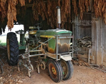 May include: A green John Deere tractor with a large rear tire, a smaller front tire, and a plow attachment. The tractor is parked in a barn with tobacco leaves hanging from the ceiling.