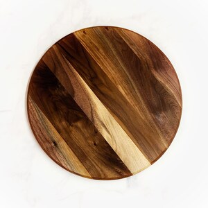 May include: A round wooden serving board made of walnut wood. The board has a natural wood finish and is perfect for serving cheese, charcuterie, or other appetizers.