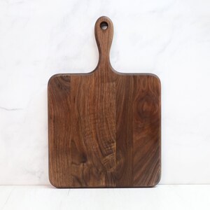May include: A square-shaped wooden cutting board with a handle. The board is made of dark brown walnut wood and has a natural wood grain pattern.
