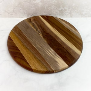 May include: A round wooden cutting board with a striped pattern of different shades of brown wood.