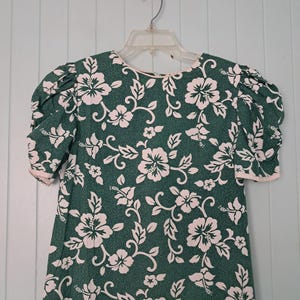 May include: A short-sleeved dress featuring a dark green background with a repeating light pink hibiscus floral pattern. The garment has puffed sleeves with light pink trim at the cuffs and a round neckline with matching light pink piping.