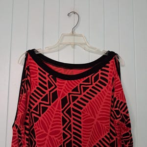 May include: Red and black patterned top with open shoulders, hanging on a clear plastic hanger. The top features a bold geometric design in black against a red background, with a black neckline and sleeve trim.