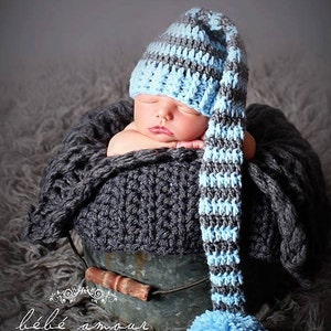 Puede incluir: Un bebé recién nacido con un gorro de punto de crochet a rayas azules y grises con una cola larga y un pompón en el extremo. El bebé está durmiendo en una manta de crochet gris dentro de un cubo de metal.