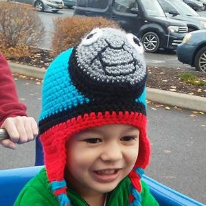 May include: A child wearing a blue, red, and gray crocheted hat with a Thomas the Tank Engine design. The hat has long braids that hang down the sides.