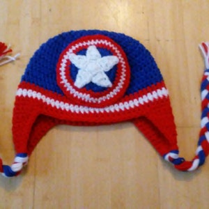 May include: A red, white, and blue crocheted hat with a star on the front. The hat has long ties that are braided.