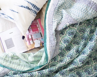 Könnte beinhalten: Eine gesteppte Tagesdecke mit einem sich wiederholenden fächerförmigen Muster in Blaugrün und Marineblau sowie einem grün gestreiften Rand. Ein offenes Buch mit dem Wort TOOLS auf der Seite liegt unter einem weißen Kissen mit blauem Batikmuster.