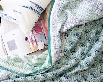 Könnte beinhalten: Eine gesteppte Tagesdecke mit einem sich wiederholenden fächerförmigen Muster in Blau und Petrol sowie einem grün gestreiften Rand, dazu ein weißes Zierkissen mit blauen Batikstreifen und ein aufgeschlagenes Buch mit dem Wort TOOLS auf der Seite.