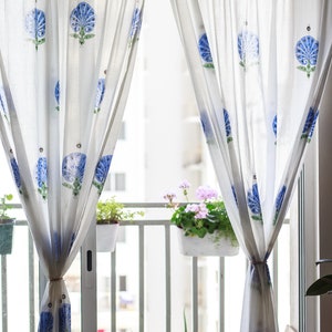 May include: White sheer curtains with a blue floral pattern hang in front of a window overlooking a balcony with potted plants.