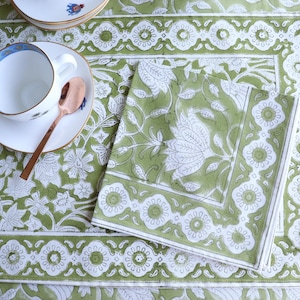 May include: A green and white floral patterned placemat, napkin, and teacup set. The teacup is white with a blue rim and sits on a matching saucer. A copper spoon rests on the saucer. The set is ideal for a tea party or dining setting.