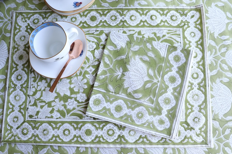 May include: A green and white floral patterned placemat, napkin, and teacup set. The teacup is white with a blue rim and sits on a matching saucer. A copper spoon rests on the saucer. The set is ideal for a tea party or dining setting.