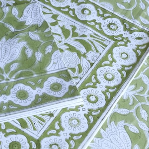 May include: A close-up of a green and white floral patterned textile. The design features large white floral and leaf motifs on a green background. A white border with floral details frames the edges. The textile appears to be folded.