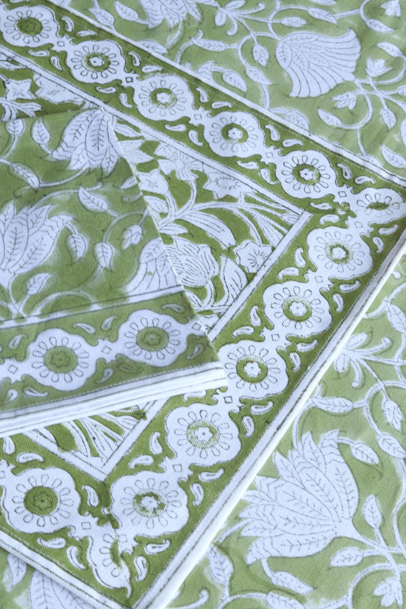 May include: A close-up of a green and white floral patterned textile. The design features large white floral and leaf motifs on a green background. A white border with floral details frames the edges. The textile appears to be folded.
