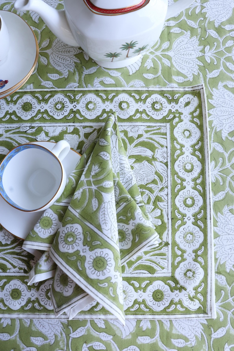 May include: A green and white floral patterned tablecloth with matching napkins and a teacup and saucer. The tablecloth and napkins feature a repeating floral design. A white teapot with a red rim is visible in the background.