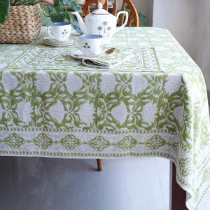 Op de afbeelding: Een groen en wit bloemen tafelkleed bedekt een houten tafel, gedekt met een theepot, kopjes en schotels. Het tafelkleed heeft een herhalend patroon van grote witte bloemen op een groene achtergrond. Een houten lepel en een gevouwen servet liggen op tafel.