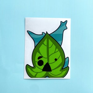 Pode incluir: Um adesivo de folha verde com um rosto de desenho animado. A folha tem um olho preto e um nariz preto. O adesivo está em um fundo branco.