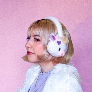 May include: A woman with blonde hair is wearing a white faux fur jacket and a pair of white earmuffs with a unicorn design. The earmuffs have purple and pink accents.