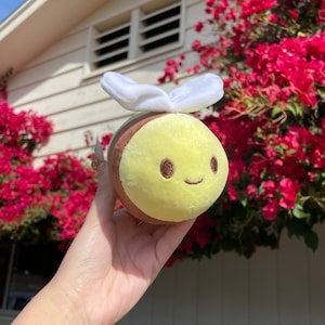 Puede incluir: Un juguete de peluche en forma de abeja con un cuerpo marrón y amarillo, alas blancas y una cara sonriente. La abeja está siendo sostenida en una mano contra un fondo de flores rosas y una casa blanca.