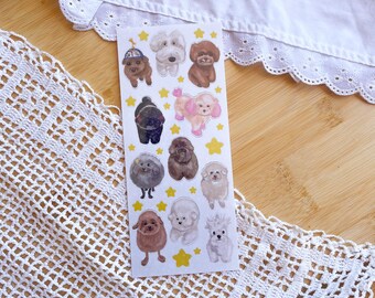May include: A sticker sheet with cartoon poodle illustrations in various colours and poses. Yellow star accents surround the stickers. The sheet rests on a wooden surface, with a white lace doily and a white fabric with a scalloped edge in the background.