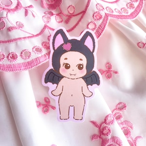 Puede incluir: Una pegatina con un bebé de dibujos animados disfrazado de murciélago. El bebé tiene una capucha negra con orejas de murciélago con un corazón rosa, alas negras y una pequeña sonrisa. La pegatina está sobre una tela bordada blanca y rosa.