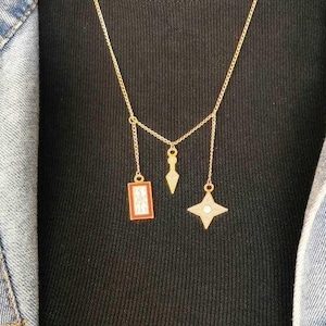 May include: A gold chain necklace with three charms: a small rectangular charm with a red and white design, a small star-shaped charm with a white center, and a small arrow-shaped charm.