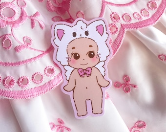 May include: A sticker featuring a cartoon baby wearing a white cat-eared hat and a pink bow tie. The baby has a smiling face and is standing. The sticker is on a pink and white fabric background with embroidered floral details.