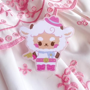 Op de afbeelding: Een gestanste sticker met een cartoon schaap verkleed als cowgirl. Het schaap draagt een roze hoed, laarzen en een gesp met een "U". Het houdt een speelgoedpistool vast. De achtergrond is witte stof met roze borduursel.