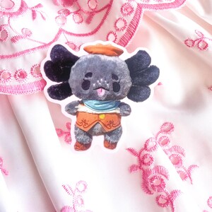 May include: A plush koala toy with a grey body, black ears, and a small orange hat. The koala wears a blue scarf and an orange waistcoat. The background is a pink fabric with embroidered floral patterns.