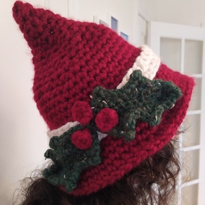Peut inclure: Un chapeau rouge en crochet avec une bande blanche et des accents de houx vert. Le chapeau a un sommet pointu et est décoré de feuilles vertes et de baies rouges.