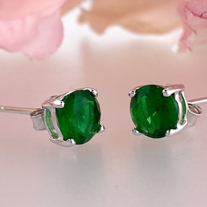 May include: A pair of stud earrings featuring round, vibrant green gemstones set in a silver metal. The earrings have a classic, minimalist design, perfect for everyday wear or special occasions.