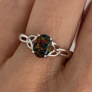 May include: A silver ring featuring an oval-shaped opal gemstone with a vibrant display of colors. The ring's band incorporates a Celtic knot design on each side of the gemstone, adding a touch of intricate detail to the jewelry.